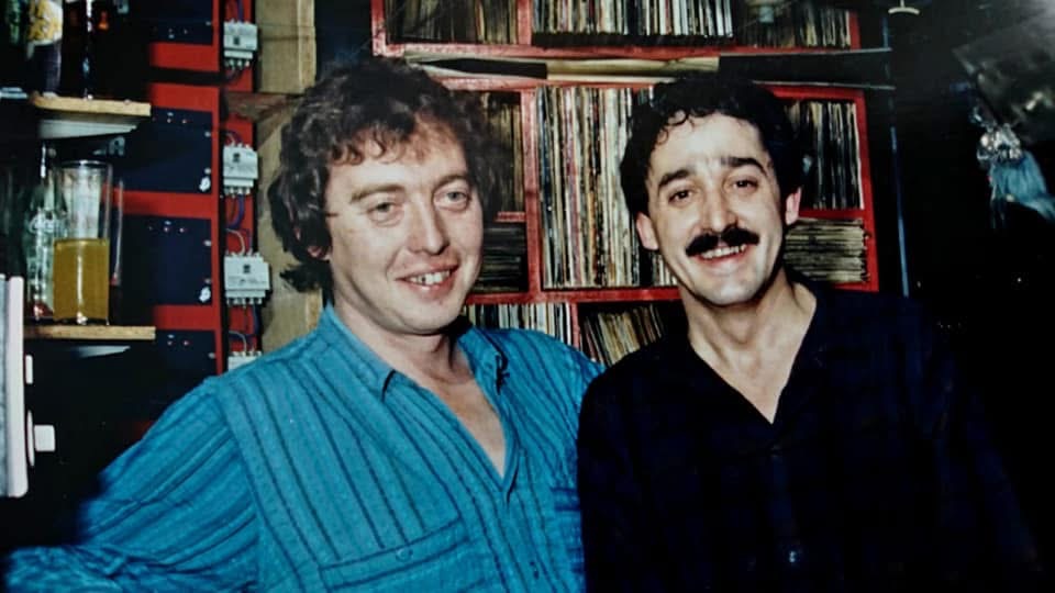 Dos disc jockeys sonriendo frente a la discoteca de vinilos